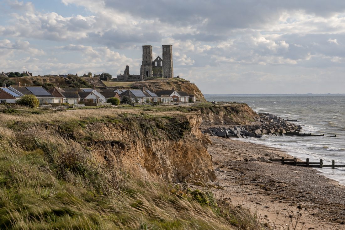 Low eroding clay clifftop with the two ruined Reculver church towers on the skyline and a row of bungalows with solar panels below them, above a shingle shore and sea defences